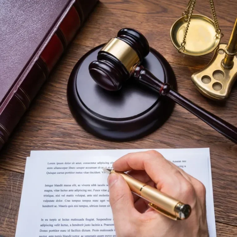 Main d'une personne écrivant avec un stylo sur un document, à côté d'un marteau de juge et de balances.