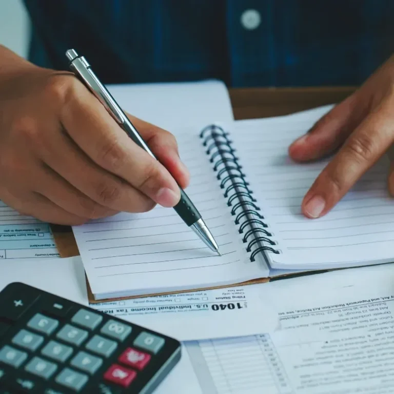 Main dans un carnet avec un stylo, une calculatrice et des documents sur la table.
