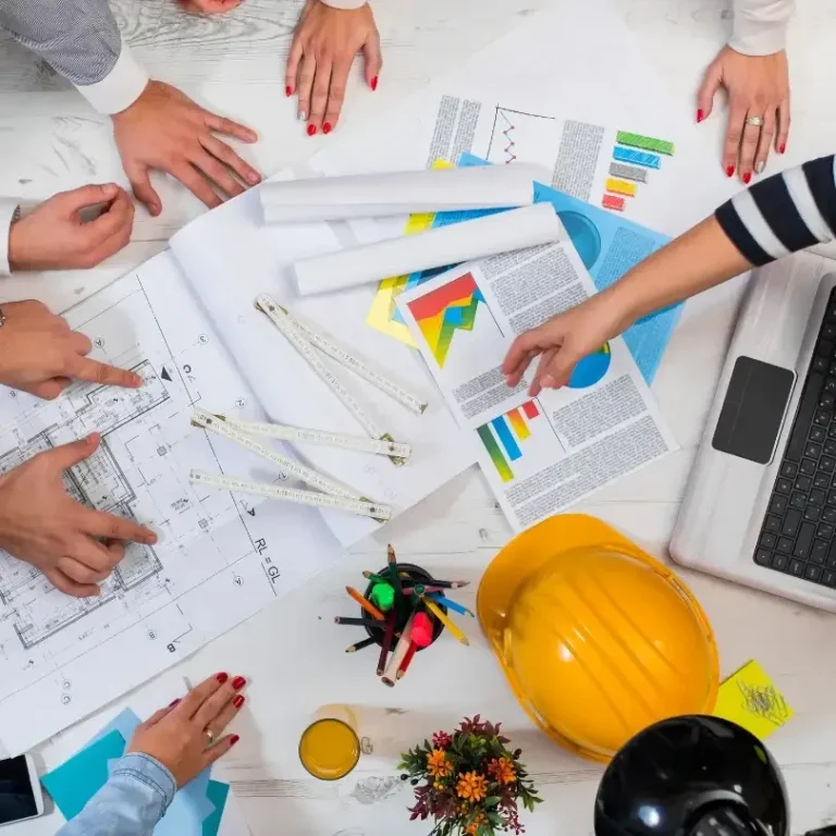 Diverses mains travaillent sur des plans et graphes, avec un ordinateur portable et un casque de chantier.